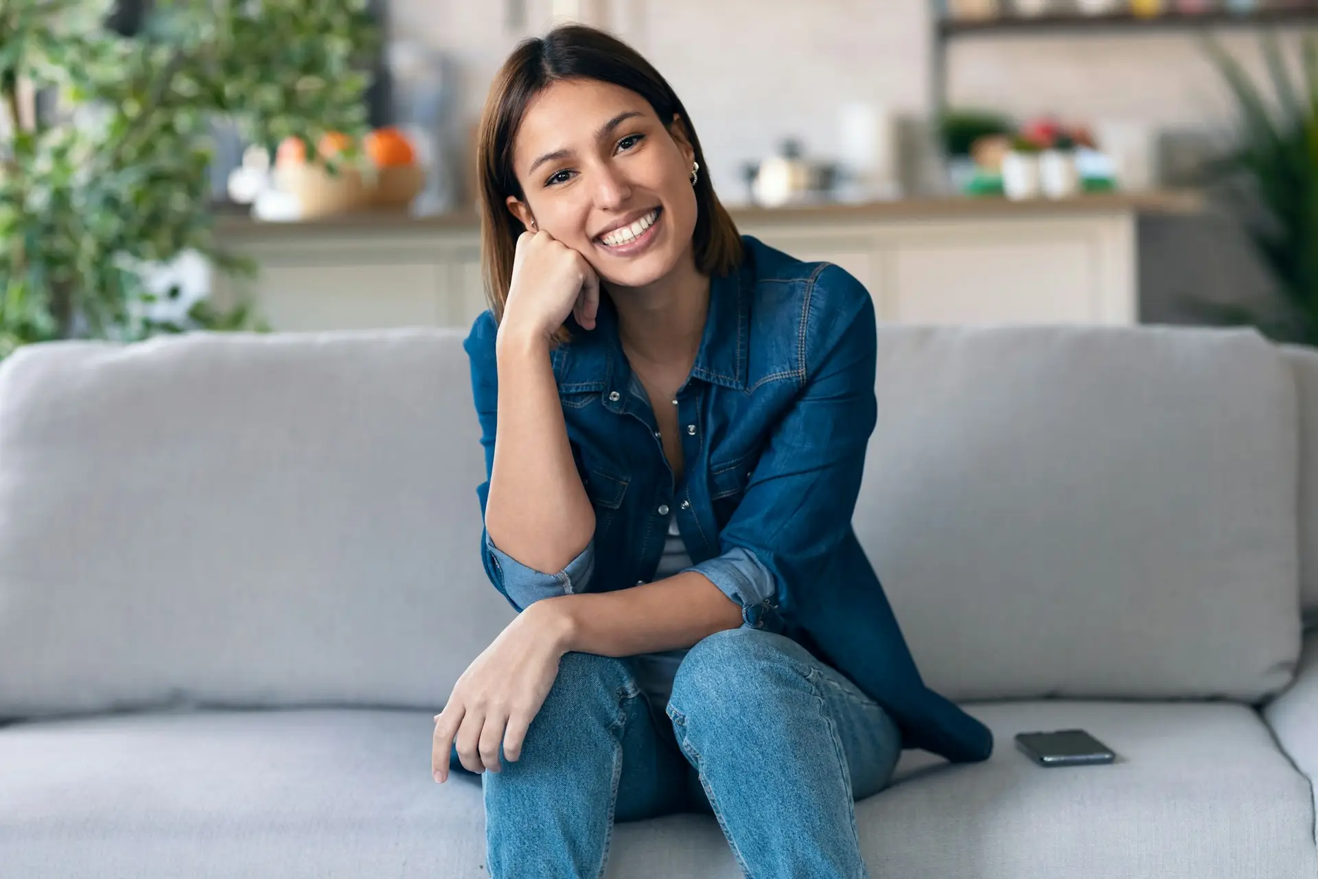 Smiling young woman looking at camera sitting on couch in the living room at home.